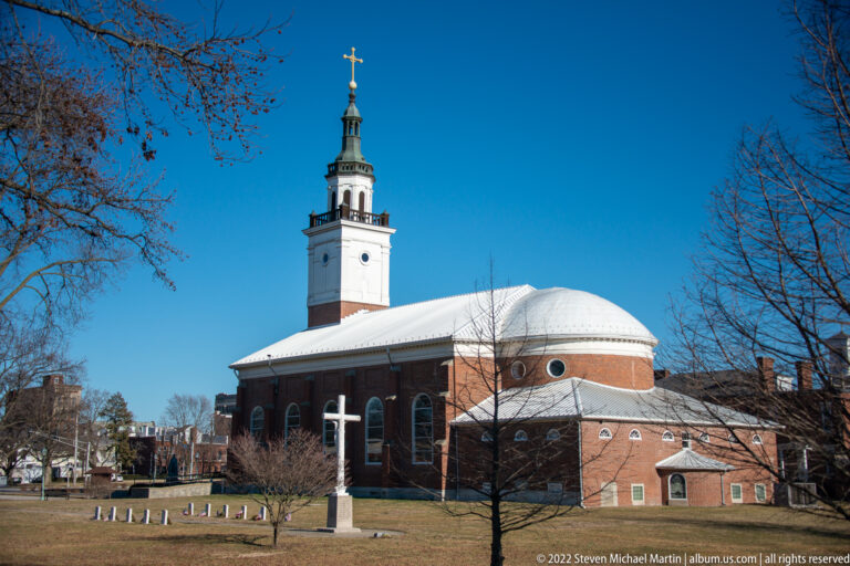 A red-brick church with a pale dome and tall bell tower stands beyond a lawn and white cross.