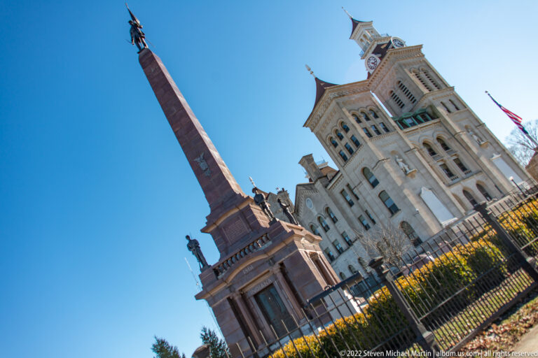 A stone obelisk stands beside a domed brick church and tower, with fencing and an American flag nearby.