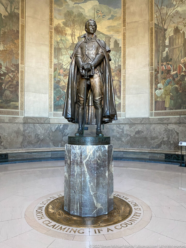 A bronze uniformed figure stands on a marble pedestal between two historical murals.