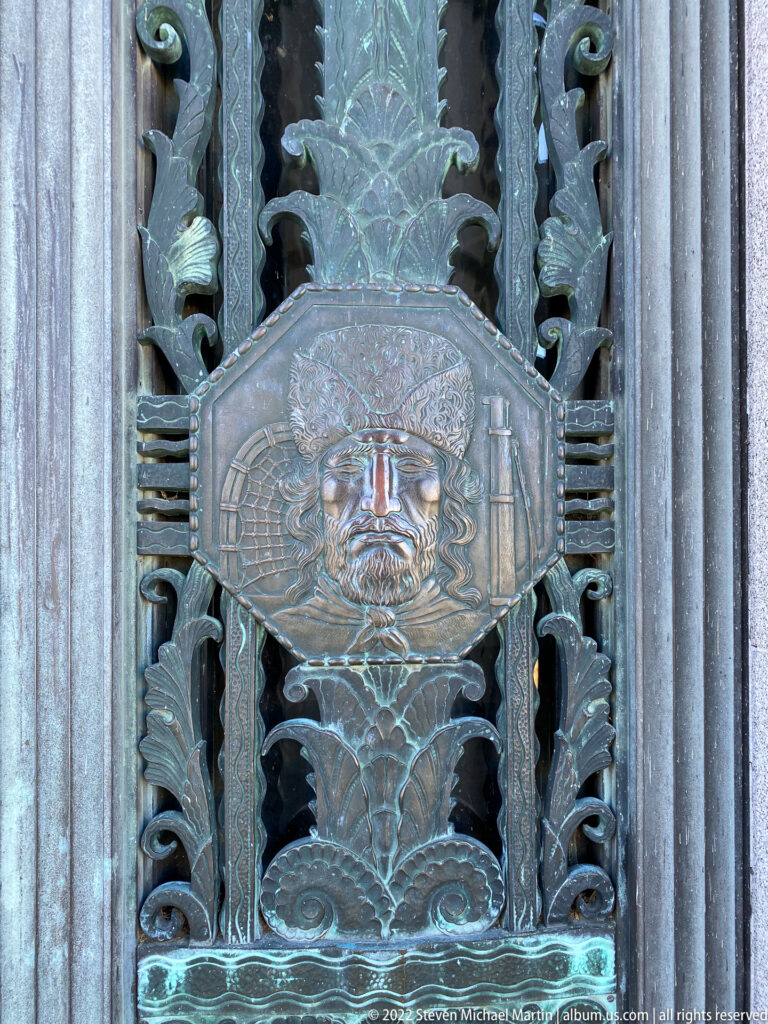 A raised portrait medallion sits amid symmetrical foliage and bands on a dark metal panel.