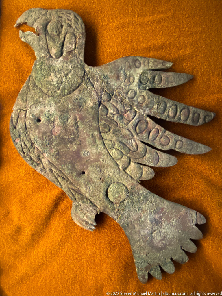 A weathered copper object with a broad body and long feather-like projections lies on orange fabric.