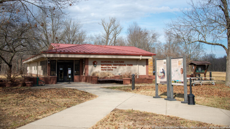 A low visitor center with a red roof stands beyond a curved path and outdoor signs.