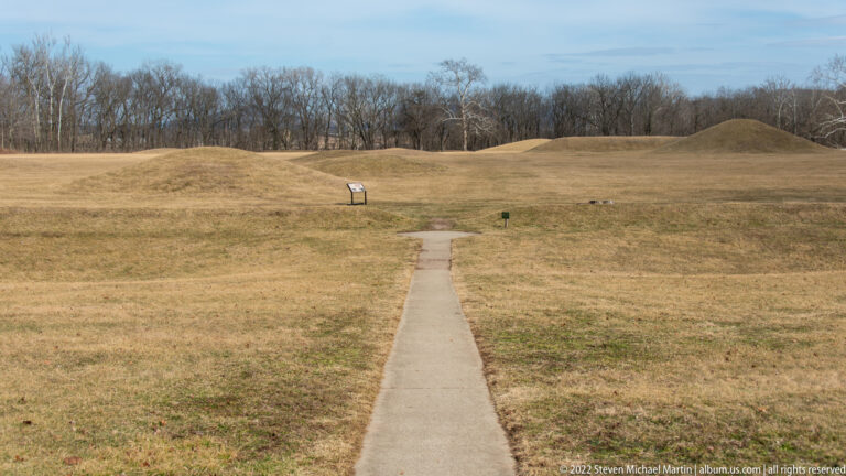 A straight concrete path crosses grass toward low mounds and an interpretive sign.