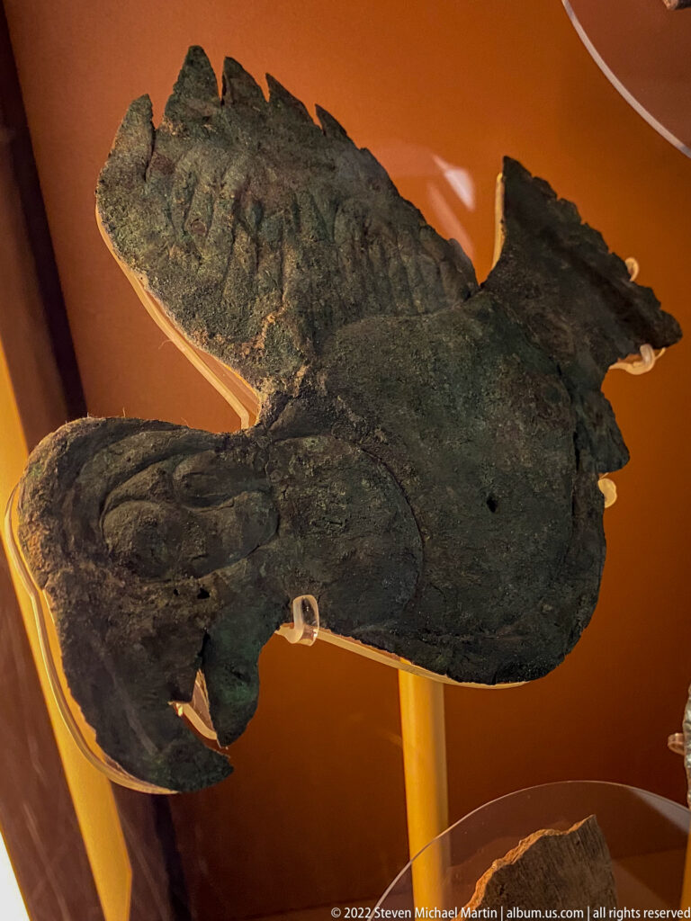 A dark copper artifact with a rounded center and irregular extended forms rests on orange backing.