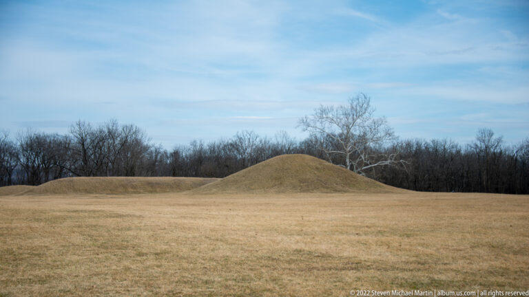 Two grassy mounds rise across an open field beneath a pale sky and bare trees.