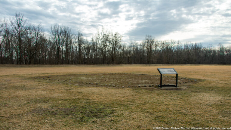 A small interpretive sign stands in a wide field before a line of leafless trees.