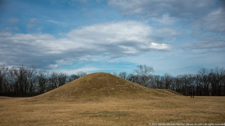 A rounded grassy mound rises above bare trees beneath a broad cloudy sky.
