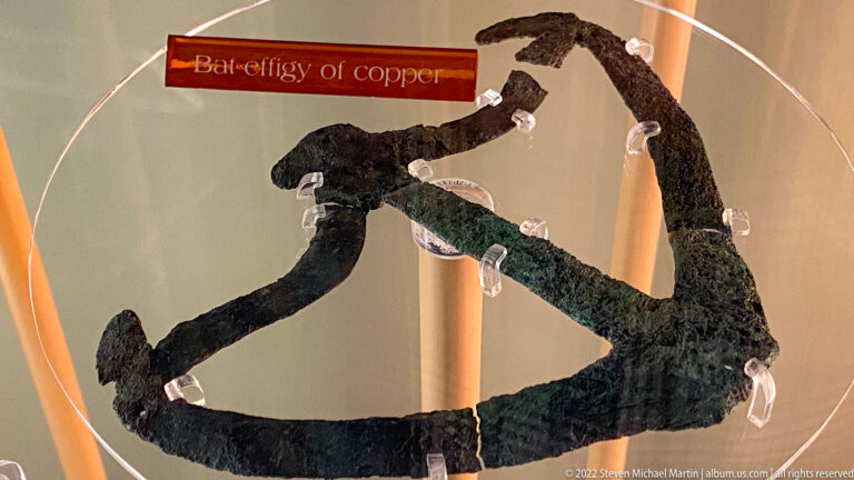 A branching, openwork copper object is held by clear supports inside a museum case.