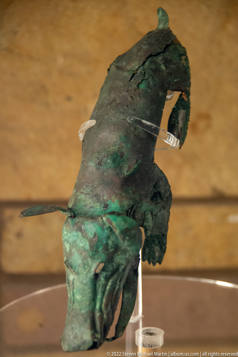 Curved green-patinated copper object with an uneven body and a narrow projection, displayed on a clear support.