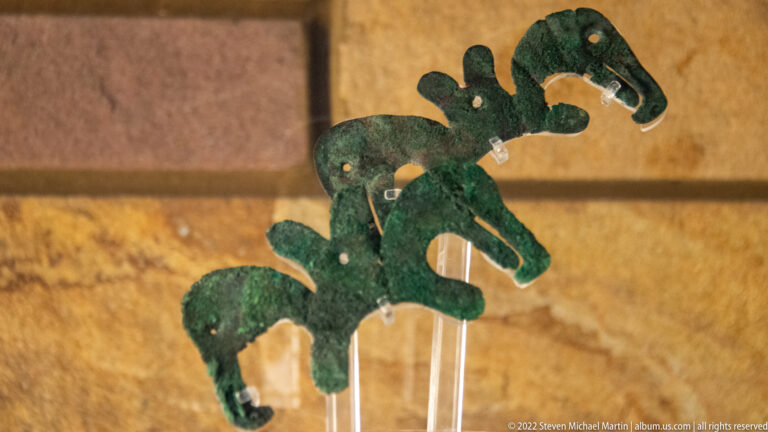 Green copper cutouts with repeated curved, birdlike outlines stand on a clear display support.
