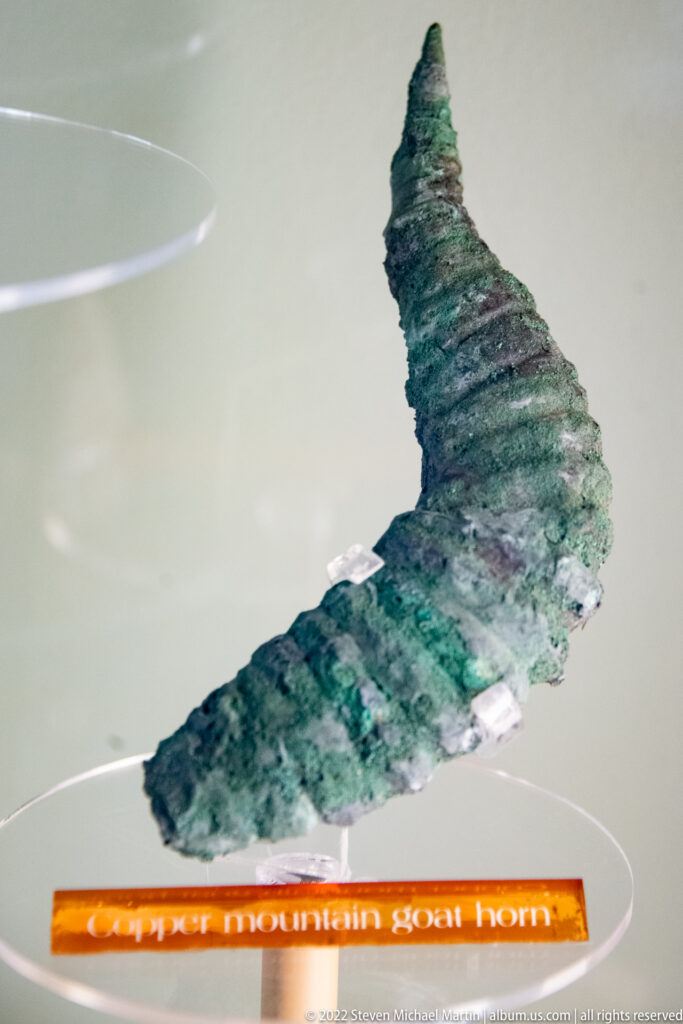 Carved green-gray effigy pipe with a long tapered form displayed above an orange label.