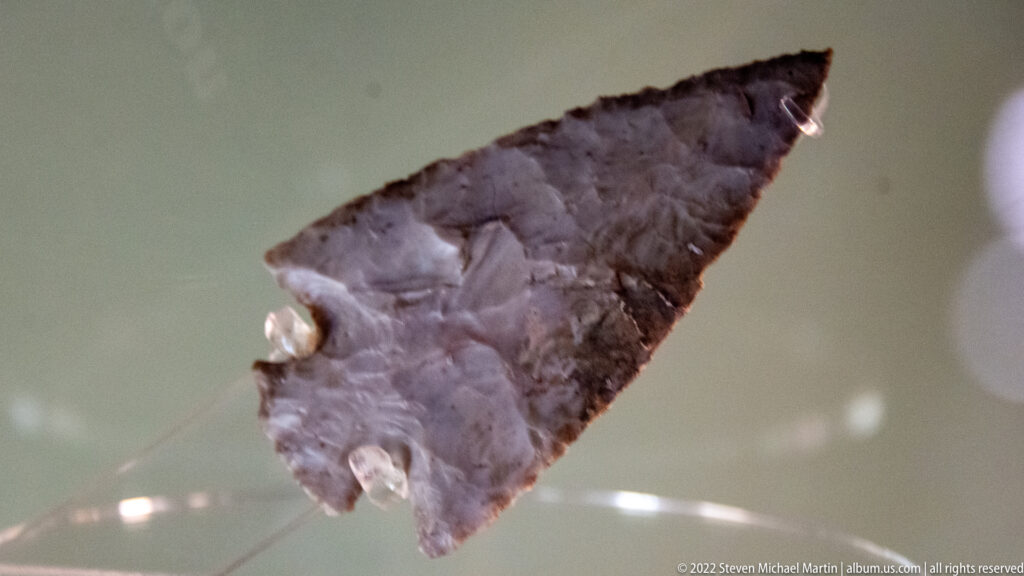 Dark triangular stone projectile point with visible flake scars on a clear museum support.