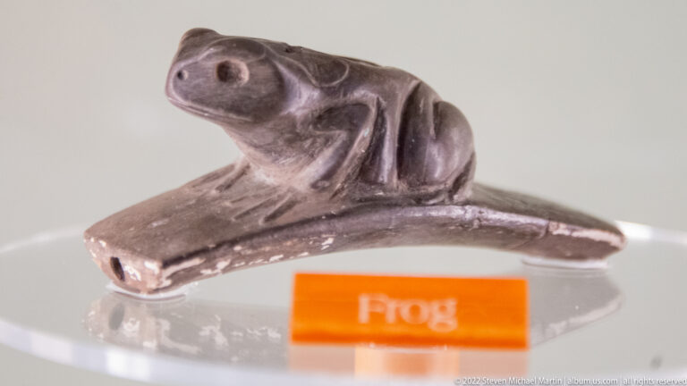 Small gray frog-shaped pipe on a flat stem, displayed above an orange label reading “Frog.”