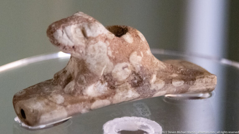 Mottled light stone pipe with a small animal carving, raised opening and flat base.