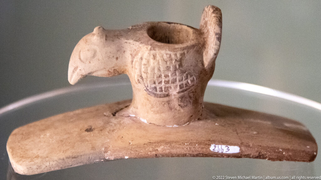Pale bird-shaped pipe with a hooked beak, patterned chest and raised opening on a flat base.