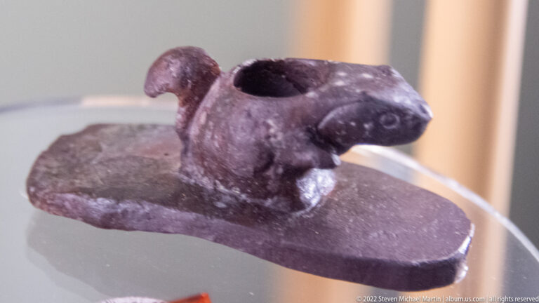 Dark stone pipe with a small animal figure, circular opening and broad base on a clear support.