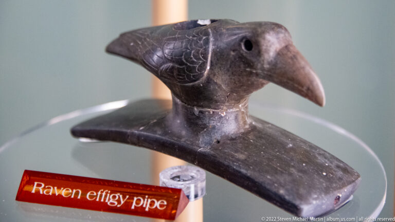 Carved raven pipe with a long beak and patterned feathers above an orange label.