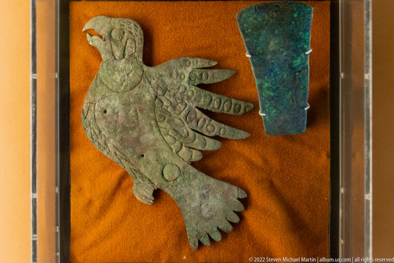 Patinated copper bird cutout lies beside a short cylindrical copper artifact in a display case.