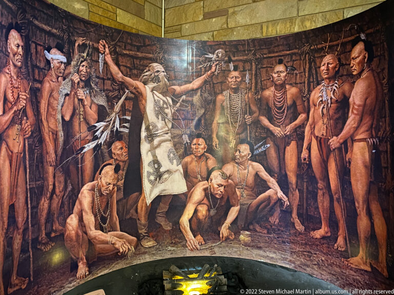 Figures gather around a fire in a museum diorama with a painted woodland background.