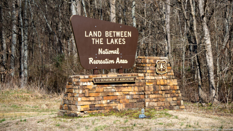 A brown National Recreation Area sign stands on a stone base beside leafless woods.