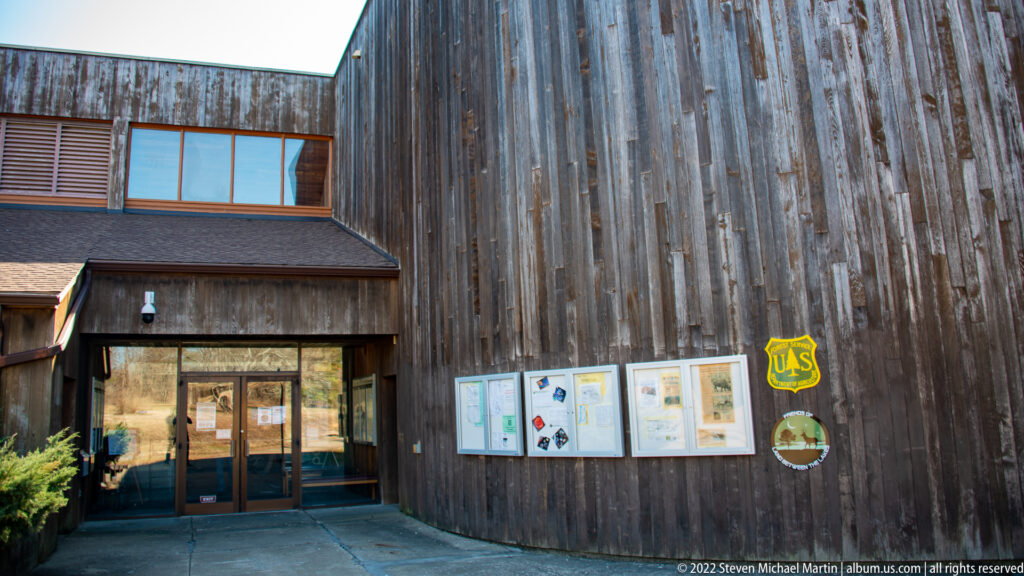 A visitor-center entrance and notice boards sit against a tall wall of weathered wood siding.
