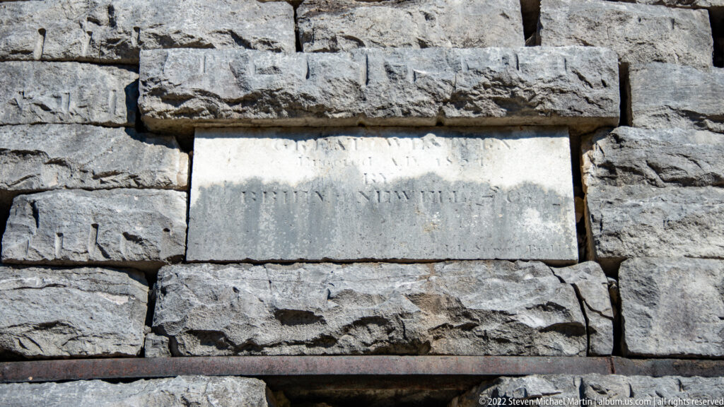 A weathered rectangular plaque sits among rough gray stones, its lettering indistinct.