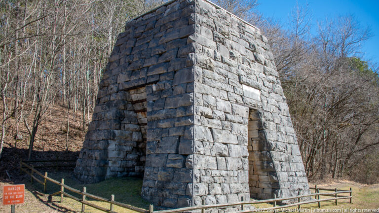 A tall stone furnace with a narrow opening stands behind a low barrier in the woods.