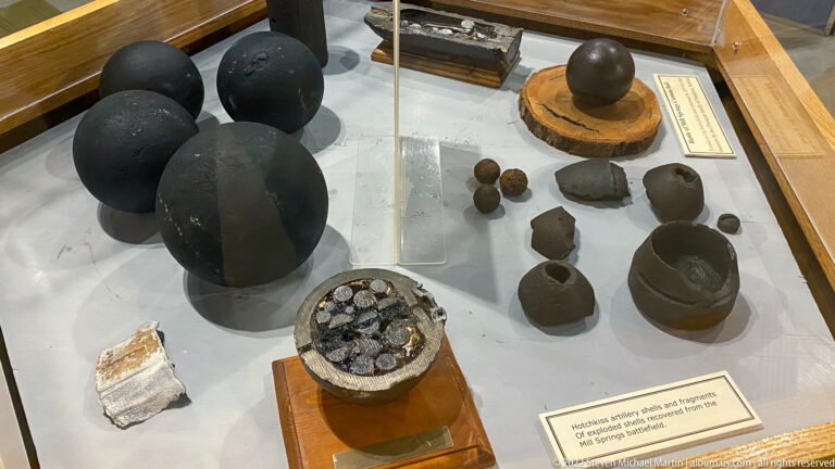 Iron cannonballs and smaller artillery fragments are arranged in a glass museum case.