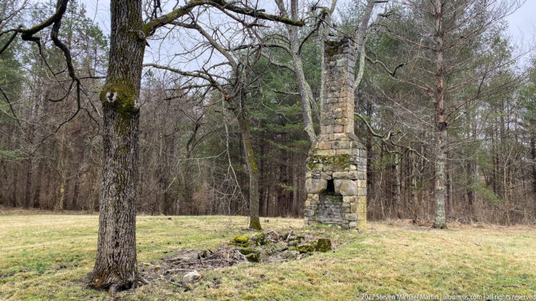A weathered stone chimney with a dark fireplace opening stands before bare trees.