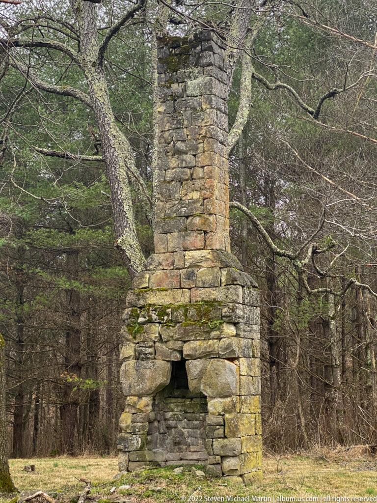 A stone chimney rises from rubble in a wooded clearing with winter grass.