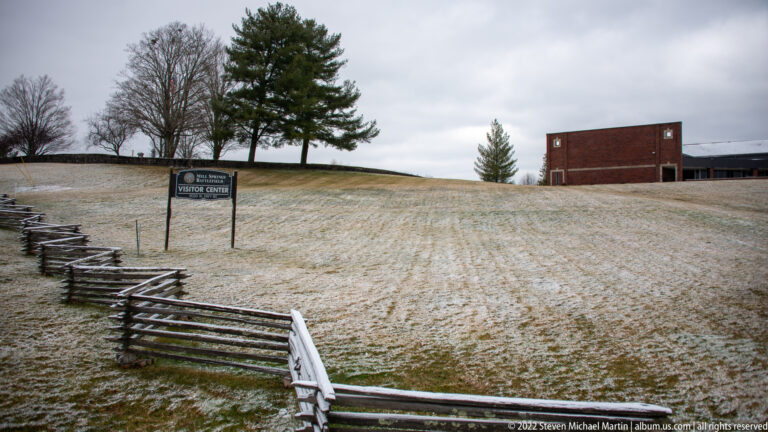 A low brick building, two flagpoles, and a cannon stand beyond a lightly snow-covered lawn.