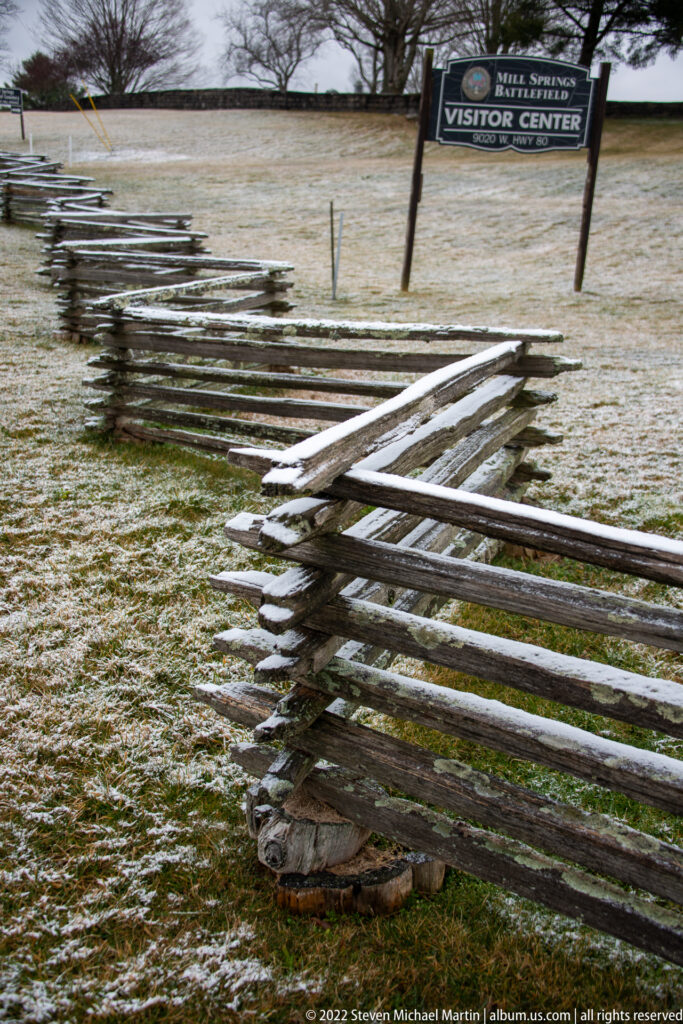 A weathered rail fence leads across snowy grass toward a visitor-center sign.