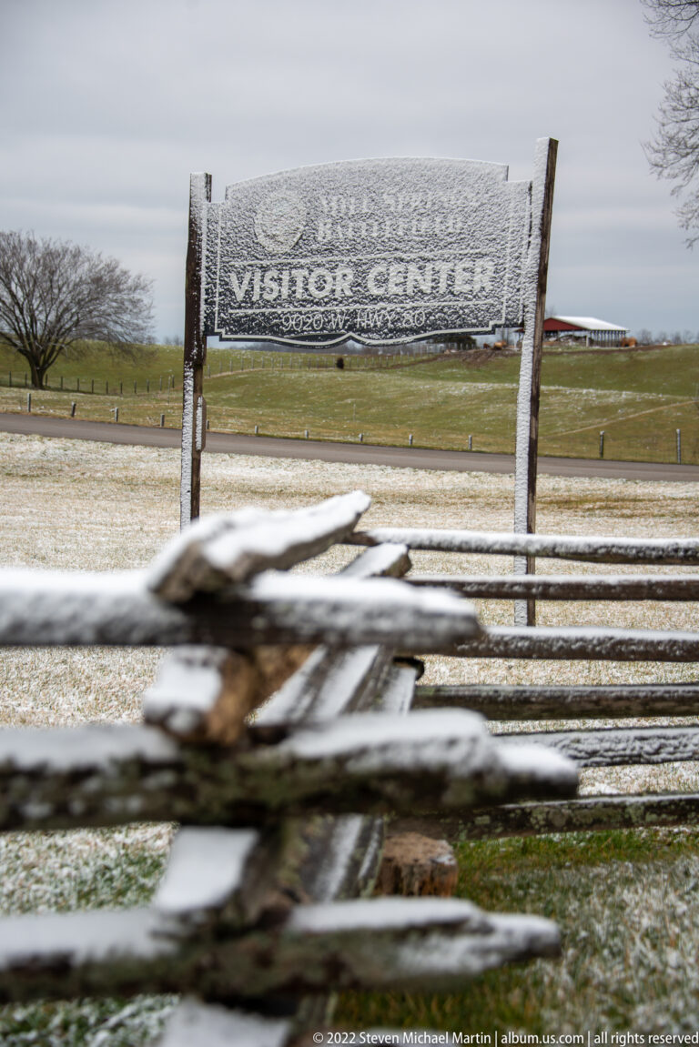 A frosted Visitor Center sign stands beyond a split-rail fence on snowy grounds.