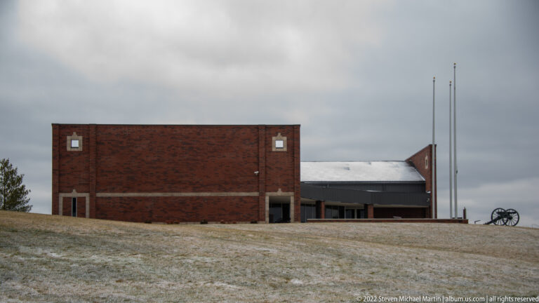A brick visitor-center building stands on a snowy rise beside flagpoles and a cannon.