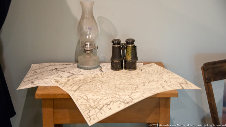 Folded map covers a wooden table beneath a glass oil lamp and two small optical instruments.