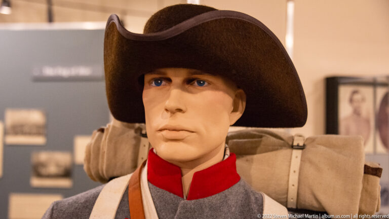 Confederate soldier mannequin in a broad hat, gray uniform and red collar, with a blanket roll behind him.