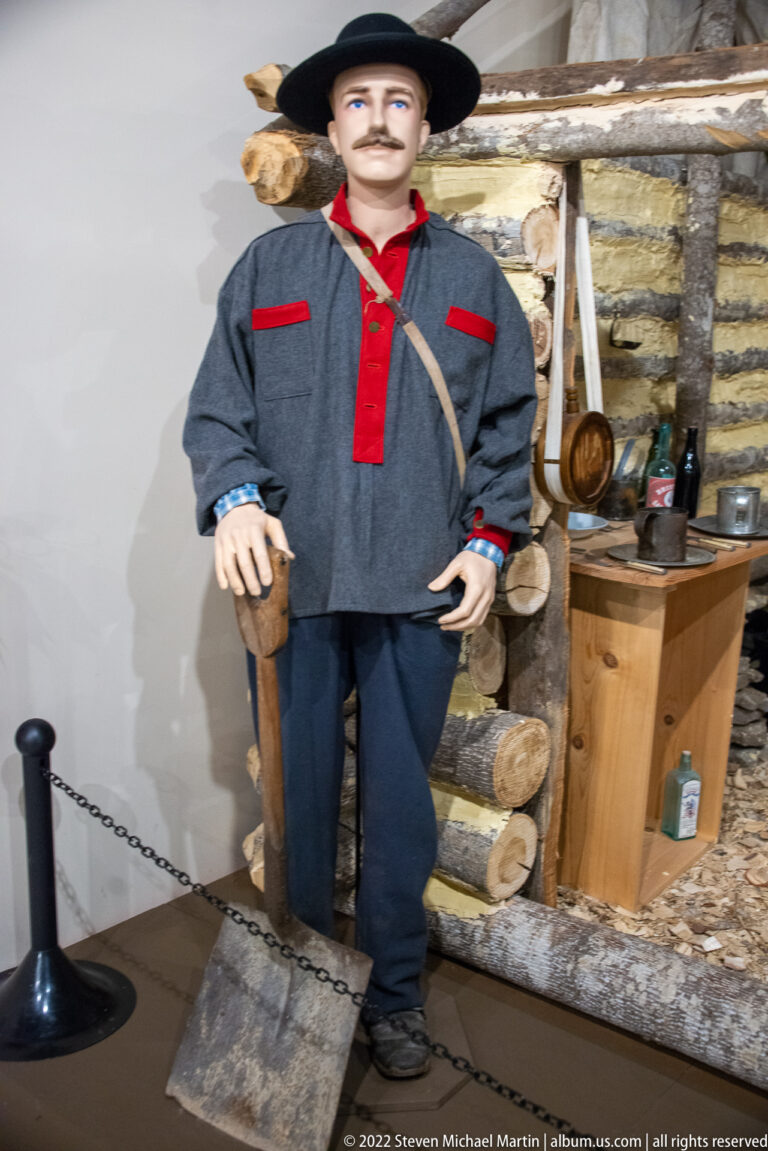 Gray-uniformed mannequin holds a long firearm beside the log wall of a cabin exhibit.