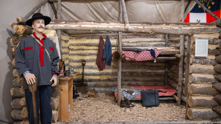 Log cabin display with a soldier mannequin, rough bunk, hanging clothes and a Confederate flag.