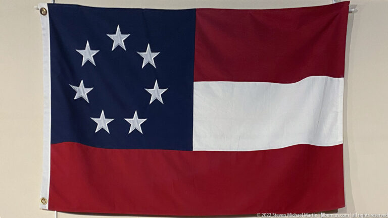 A flag with red and white stripes has a blue canton containing a circle of white stars.