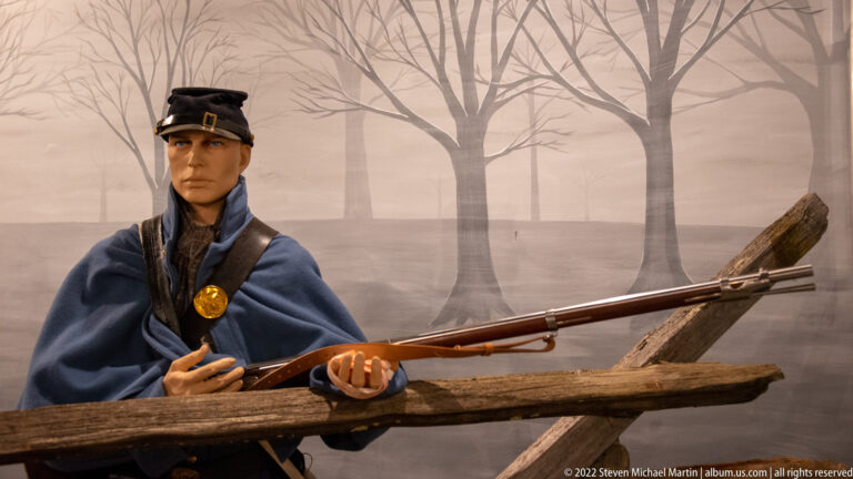 Blue-uniformed mannequin holds a rifle behind a fallen log in a painted woodland setting.