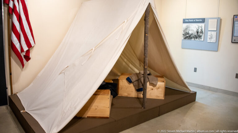 Canvas tent on a museum platform with wooden boxes, a U.S. flag and an interpretive panel behind it.