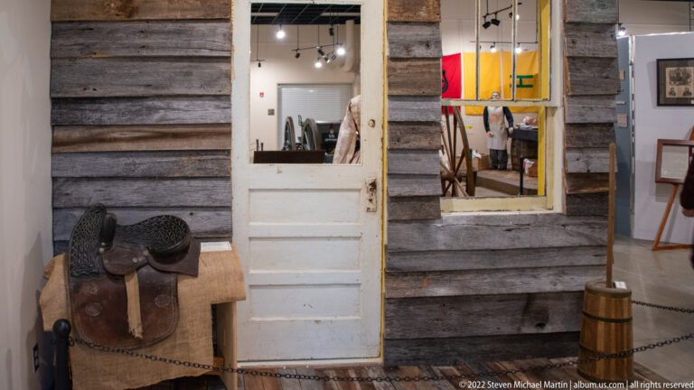 Log cabin wall and open white door frame an exhibit room, with a saddle and barrel in front.