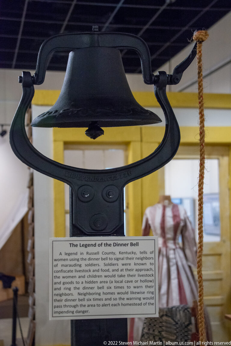 Black metal bell hangs from a curved support above a label titled “The Legend of the Dinner Bell.”