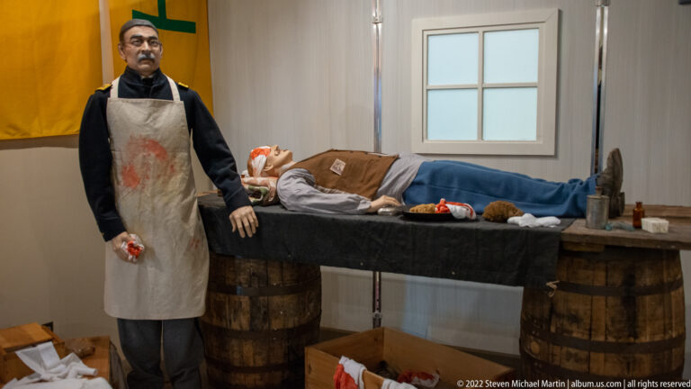 Aproned attendant stands beside a wounded patient on a plank table in a medical exhibit.