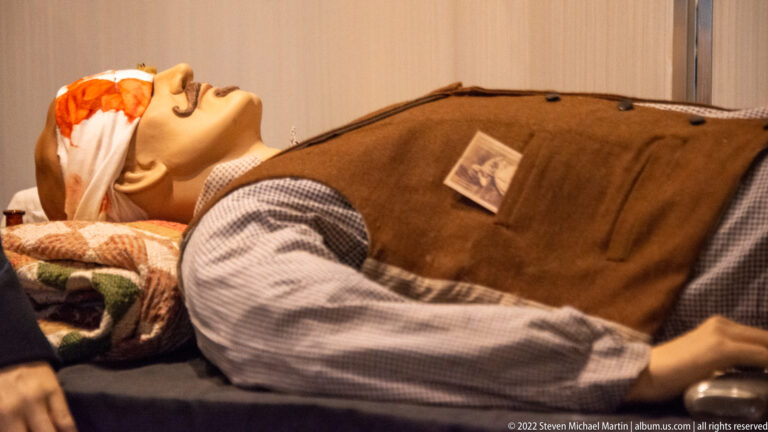 Reclining mannequin under a brown blanket, with a checked sleeve and outstretched arm visible.