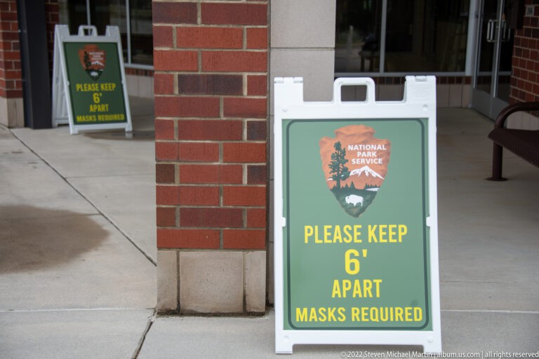 A National Park Service sign asks visitors to keep six feet apart and wear masks beside a brick visitor-center entrance.