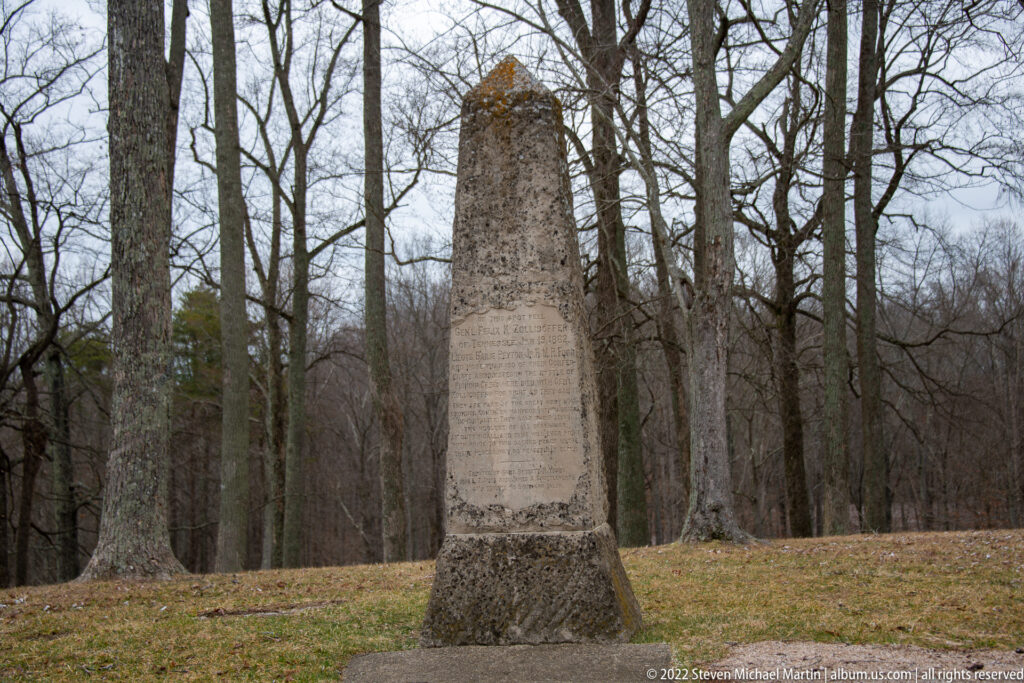 A weathered stone obelisk with an inscribed face stands on a grassy rise among leafless trees.