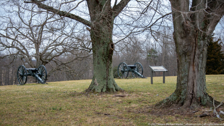 Two black field cannons stand among leafless trees on a grassy rise beside an interpretive sign.