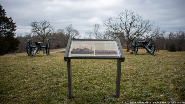 Two field cannons stand on either side of an interpretive panel on a winter lawn.