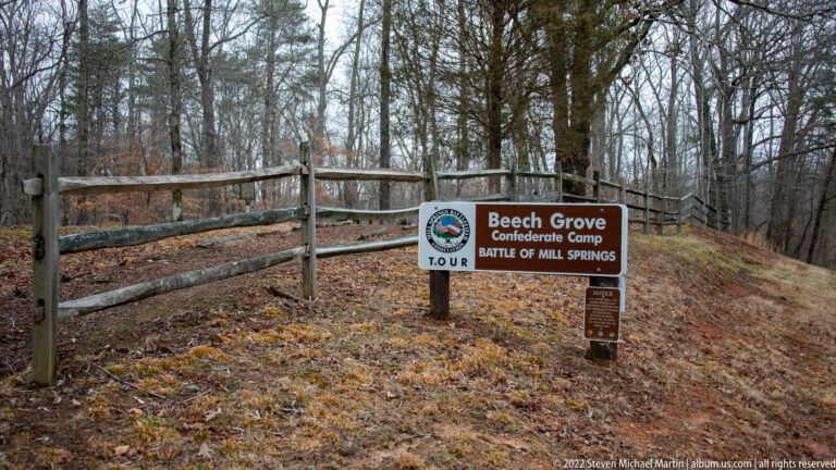 A Beech Grove Confederate Camp tour sign stands beside a split-rail fence in leaf-covered woods.
